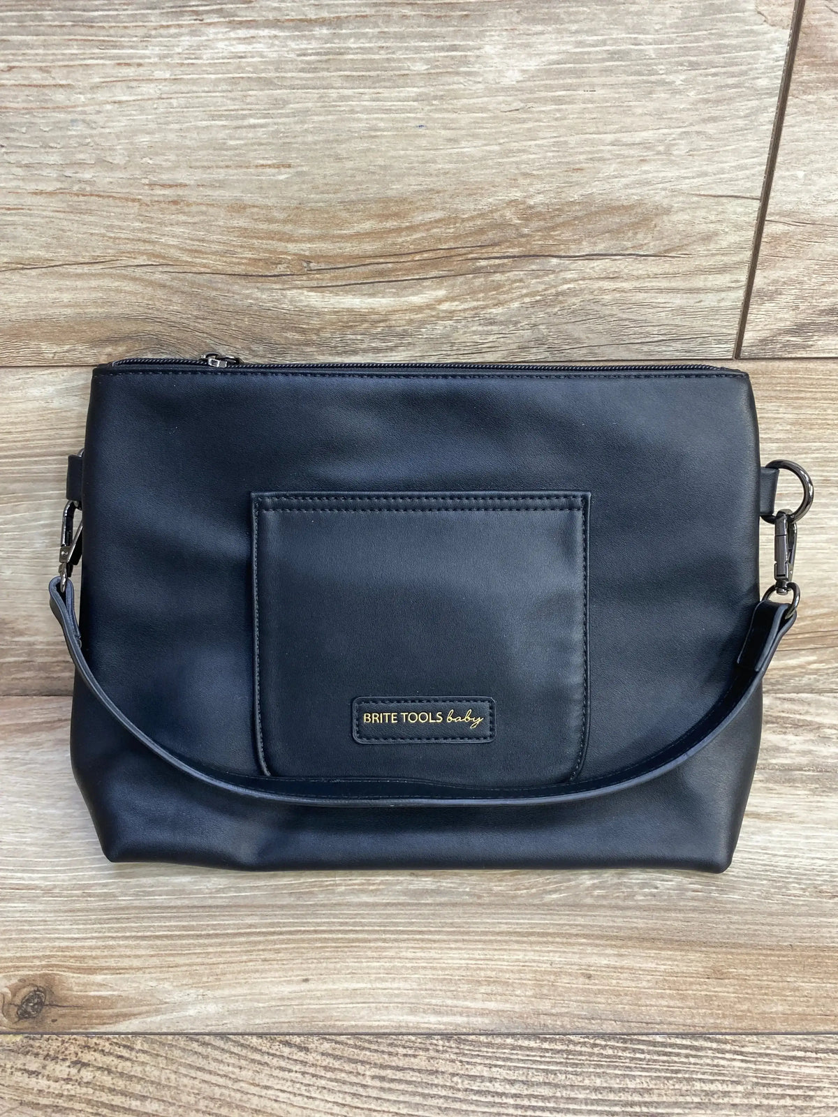 Front view of Brite Tools diaper clutch in black with front pocket, gold logo patch, top zipper and detachable wrist strap on wood