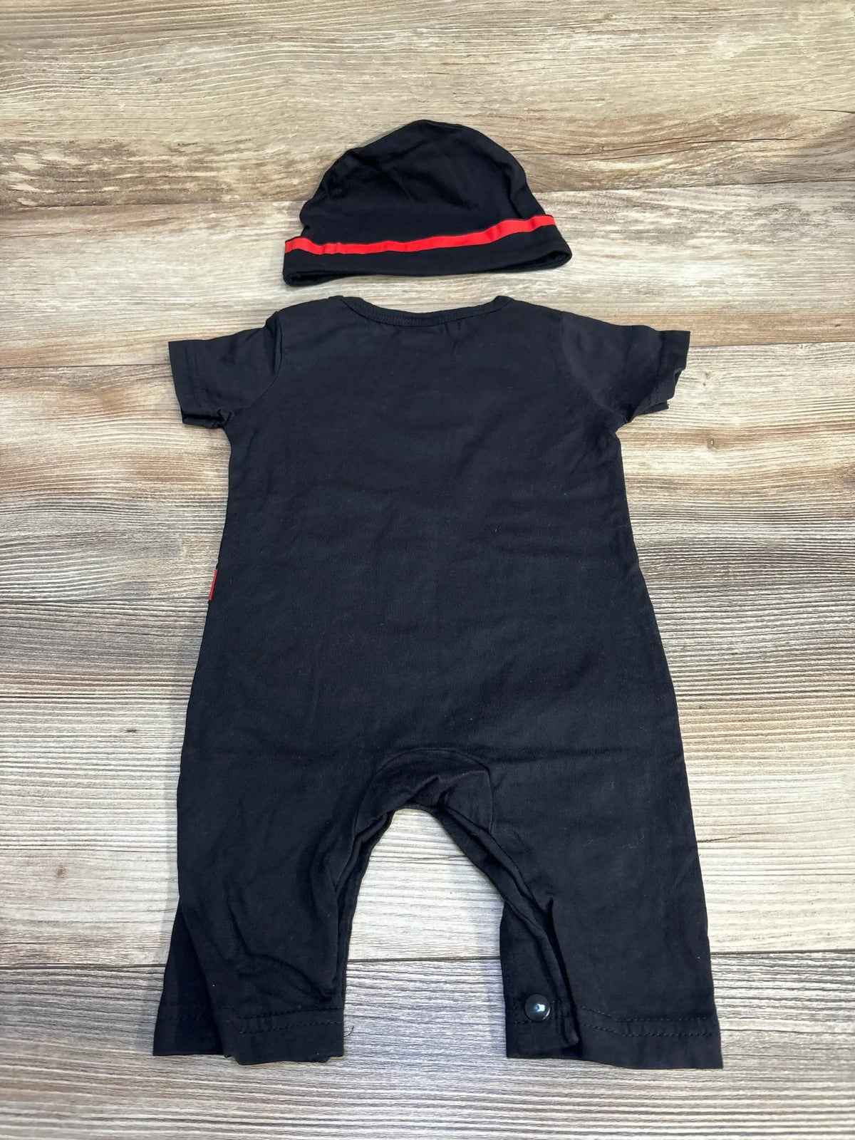 Back view of black 2-piece ninja romper and matching hat showing short sleeves, crotch snaps and red hat stripe on wood background