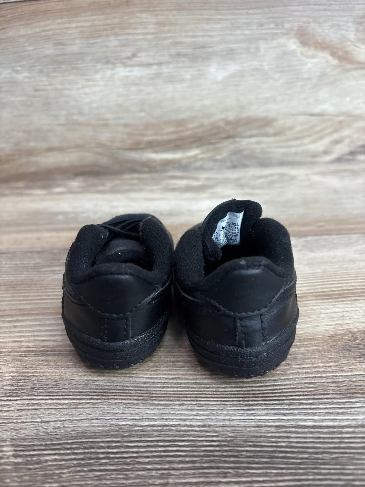 Rear view of Nike Air Force 1 crib shoes in black showing padded collar and leather upper on wood background