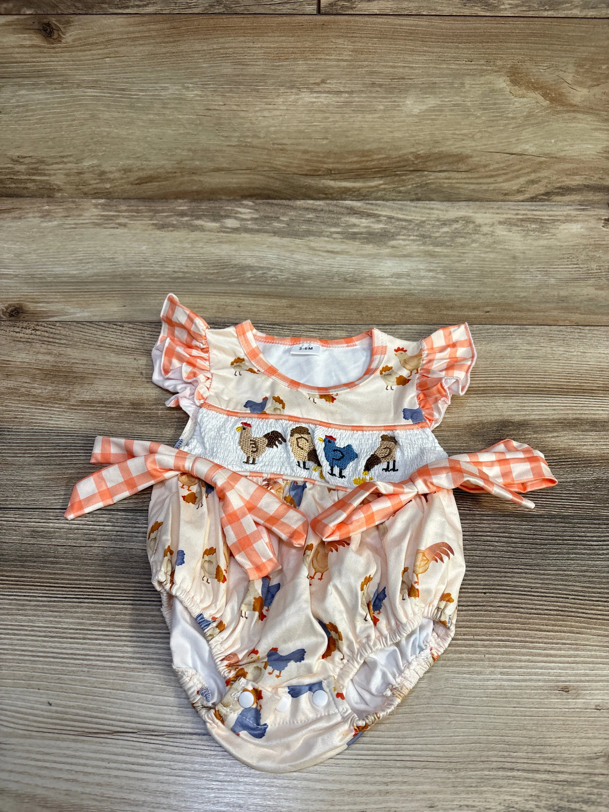 Flat lay of ivory baby bubble romper 3-6M with smocked chicken embroidery, gingham sash, ruffled sleeves on wood background