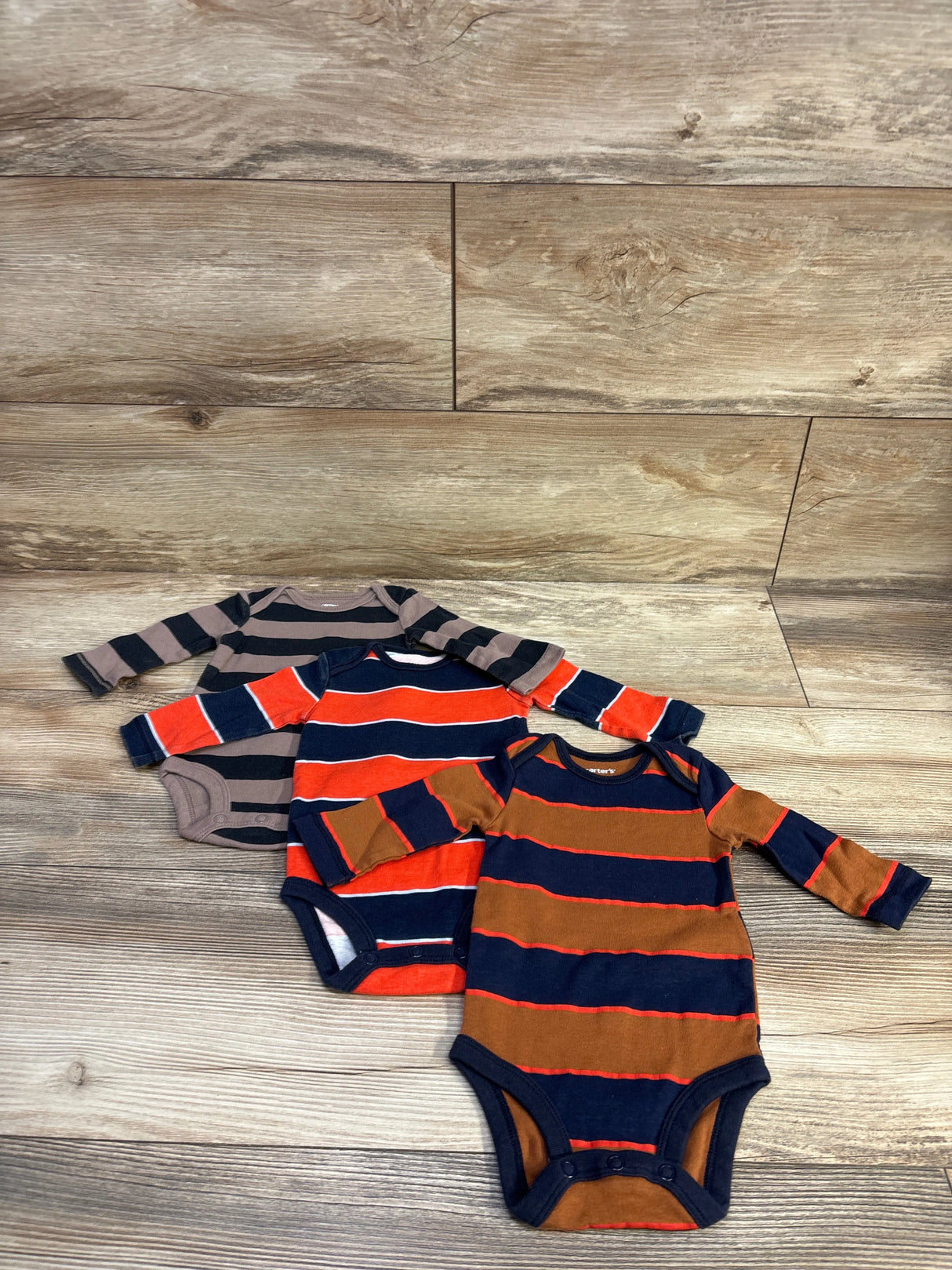 Flat lay of three Carter’s striped long-sleeve bodysuits showcasing stripe patterns and crotch snaps with wood floor background