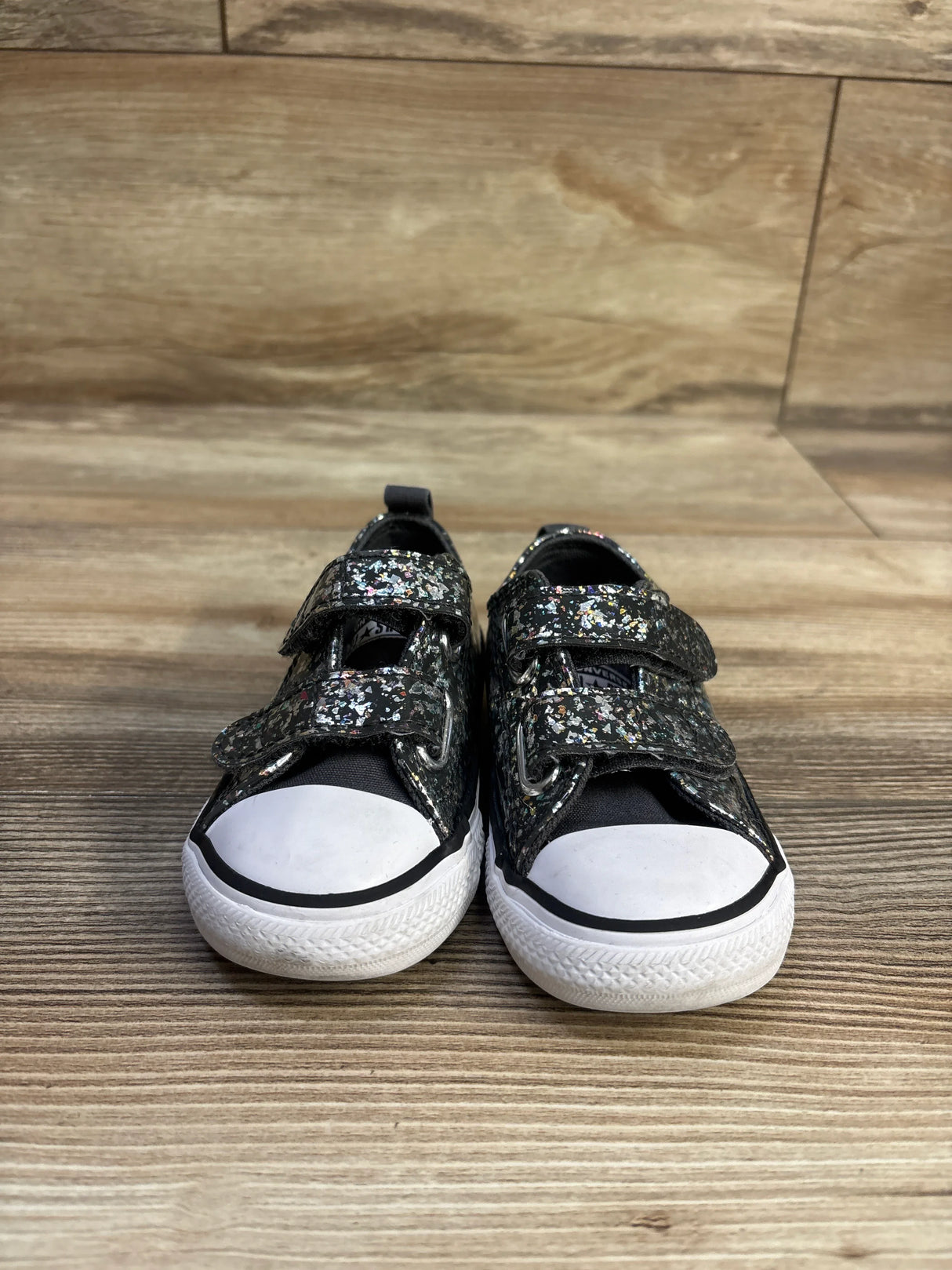 Front view of Converse Chuck Taylor sneakers size 6C with black glitter upper, Velcro straps, white rubber toe caps on wood