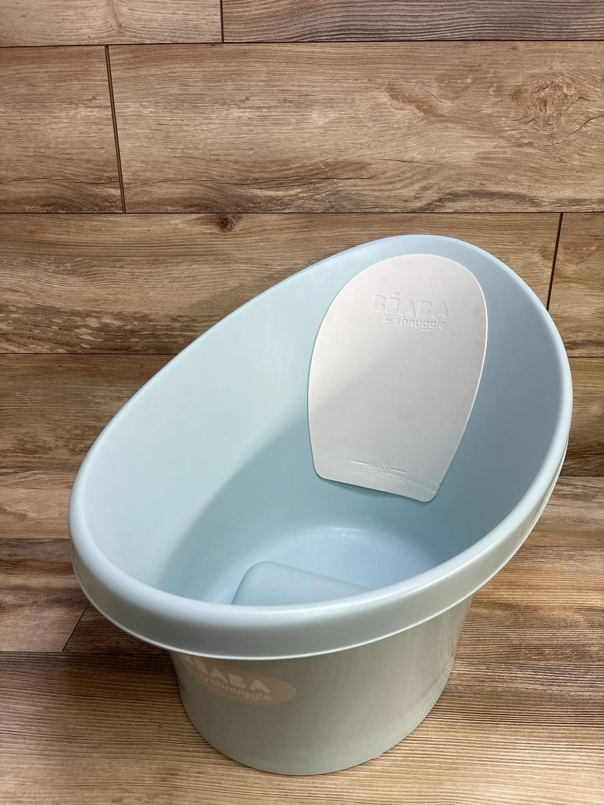Angled top view of BEABA by Shnuggle blue infant bathtub showing contoured seat and white non-slip backrest on wood floor