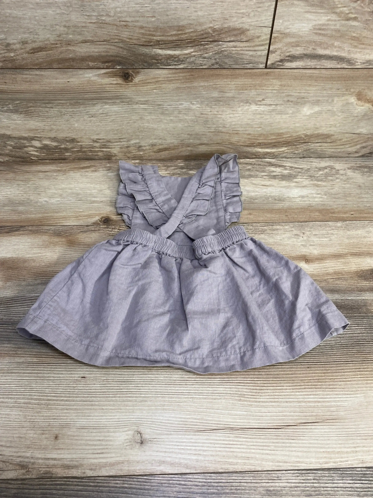 Flat lay of grey linen pinafore dress showcasing ruffled crisscross straps and elasticized waist on wood background