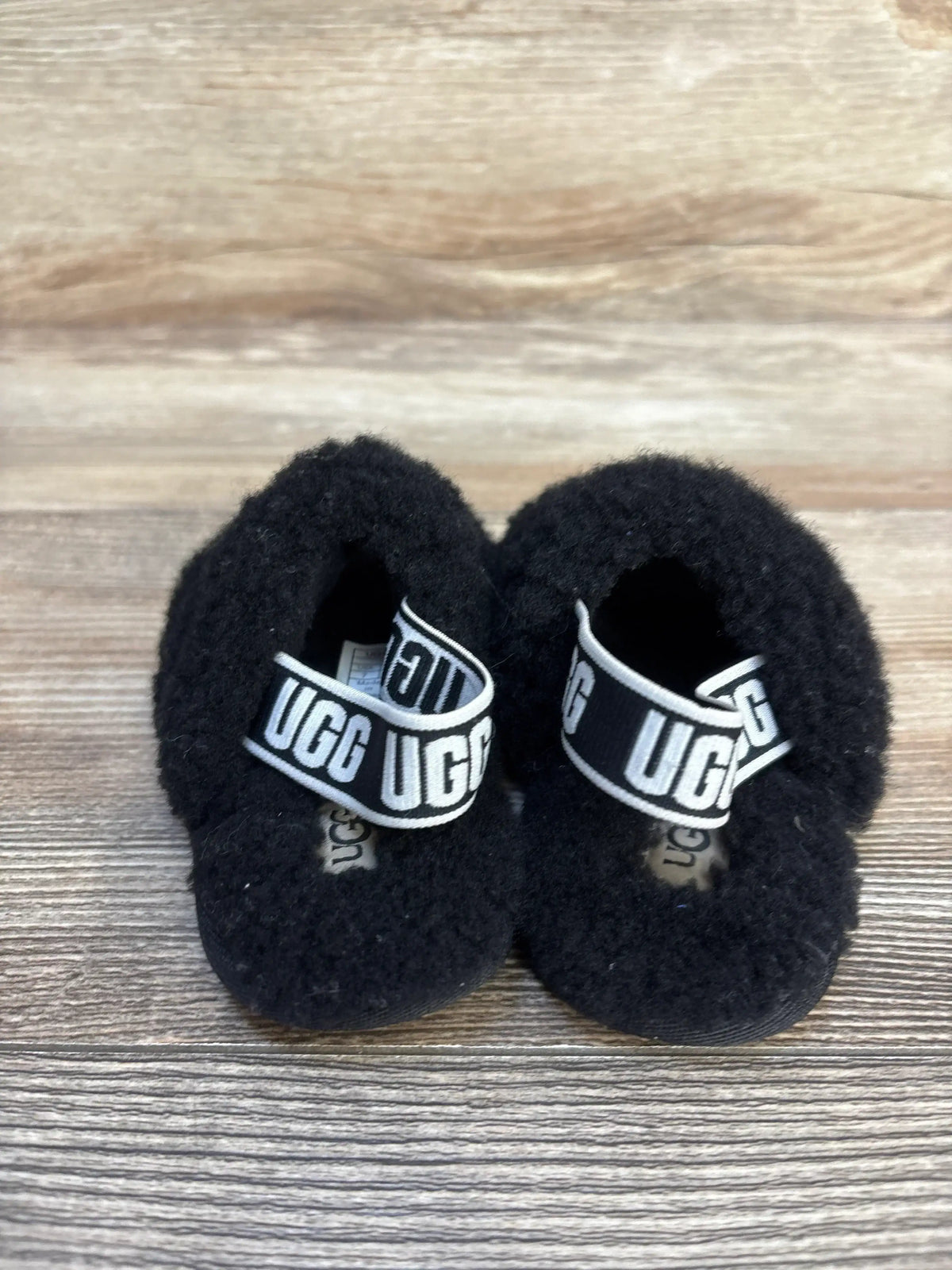 Black infant UGG fuzzy slippers with logo strap on wood background