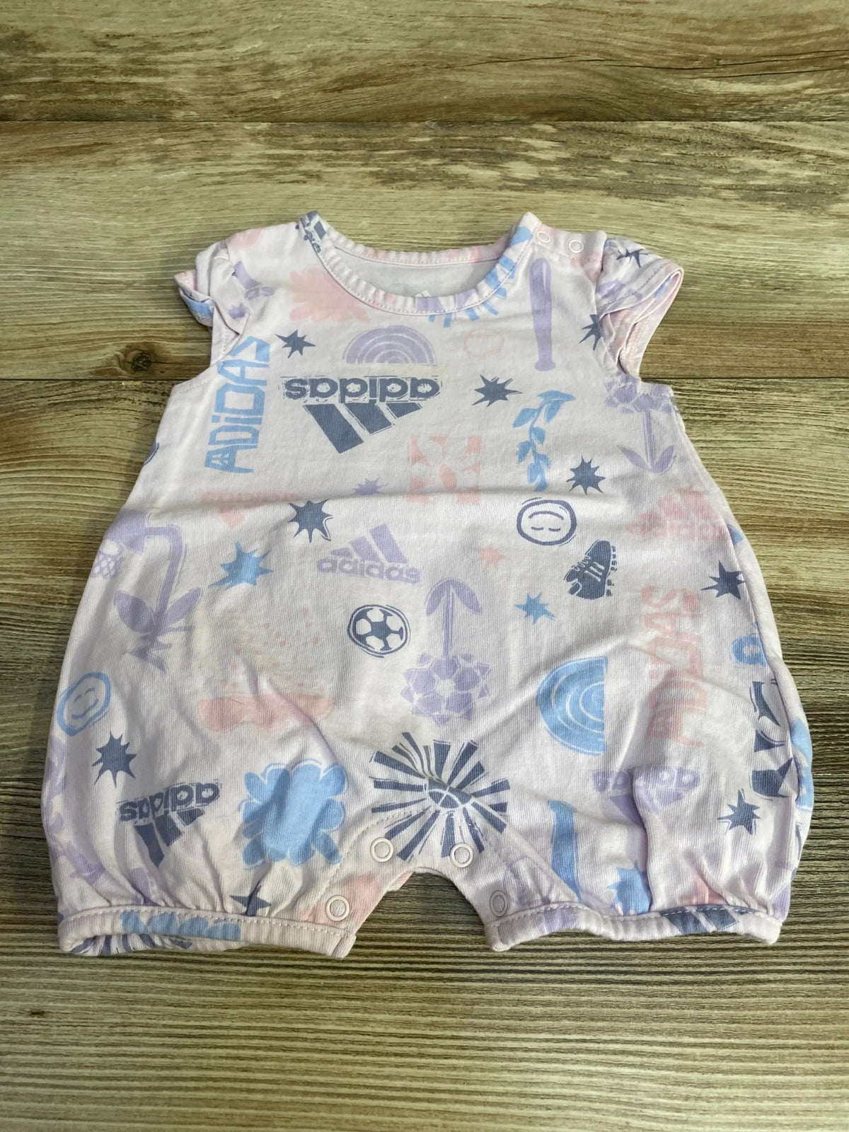 Top-down view of pink Adidas shortie romper showing all-over logo and sports graphics, snap crotch closures, laid flat on wood