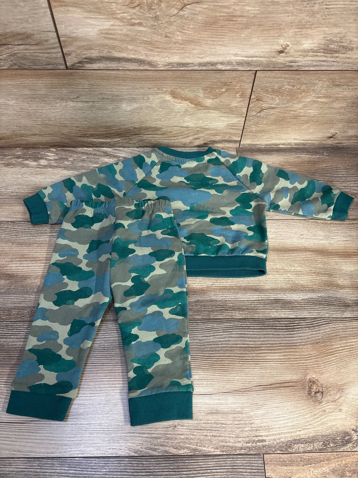 Top-down view of Cat and Jack 2-piece camo sweatshirt and joggers set in green, showcasing ribbed cuffs and waistband on a wood floor