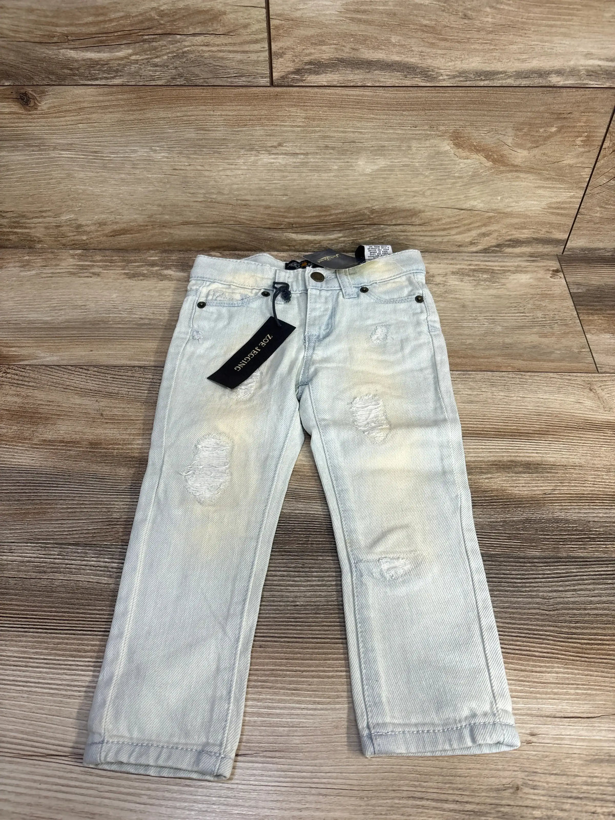 Front view of Lucky Brand Zoe toddler jeggings, light blue 2T, showing distressed knees and hang tag on wood floor
