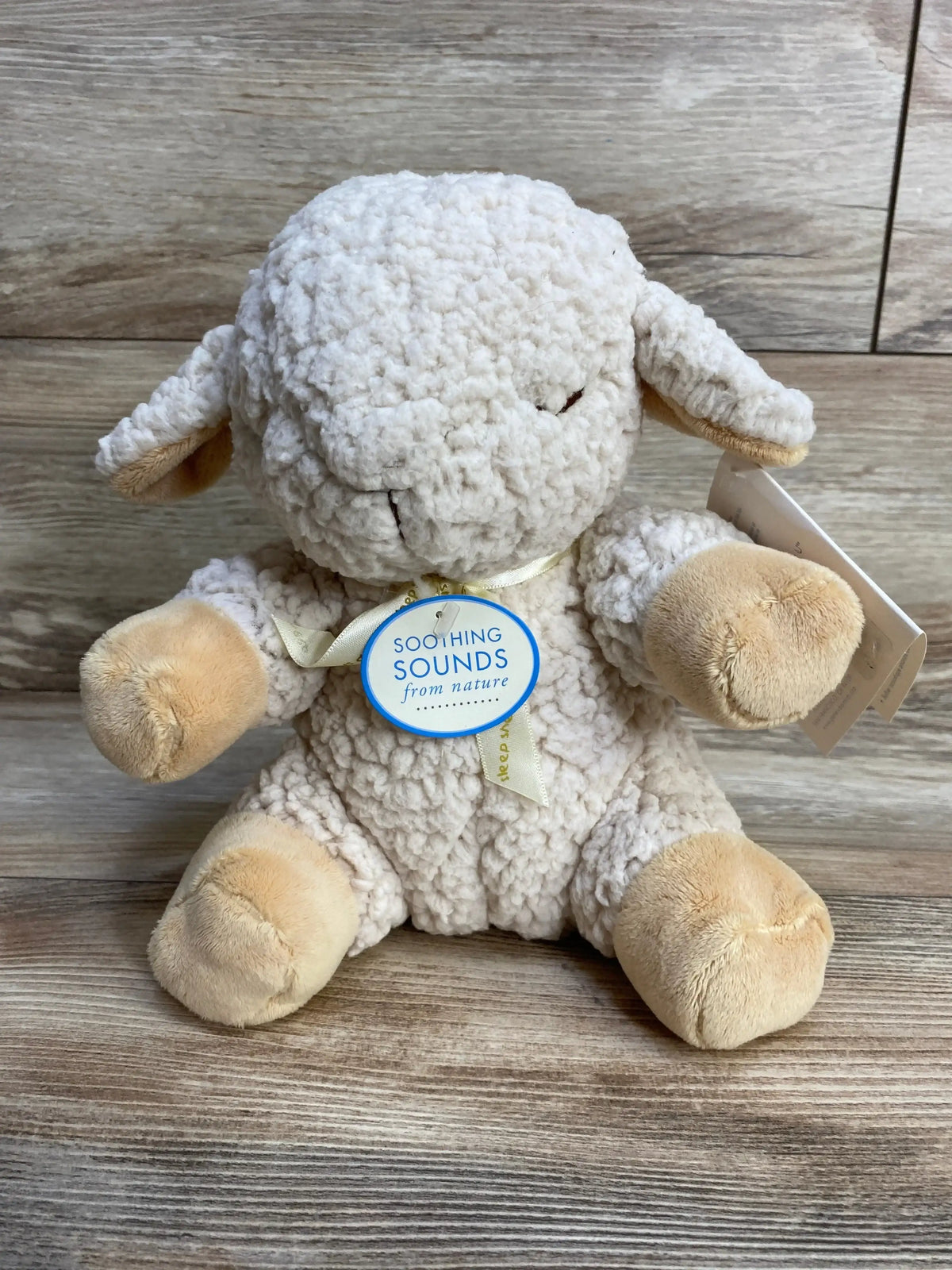 Front view of Cloud B Sleep Sheep plush showing curly ivory fleece and beige paws with round Soothing Sounds from nature tag on wood background