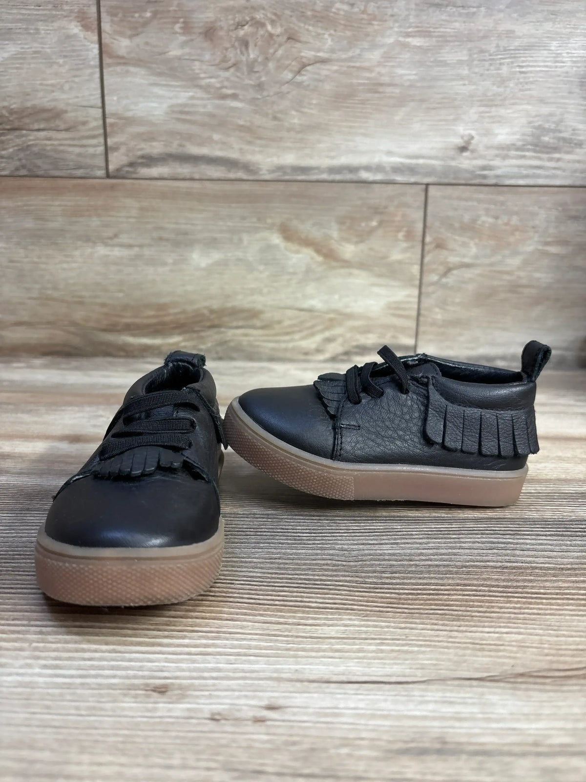 Side view of Freshly Picked Ebony Mocc Sneakers sz 6c in black leather with fringe tongue and gum soles on wood plank floor