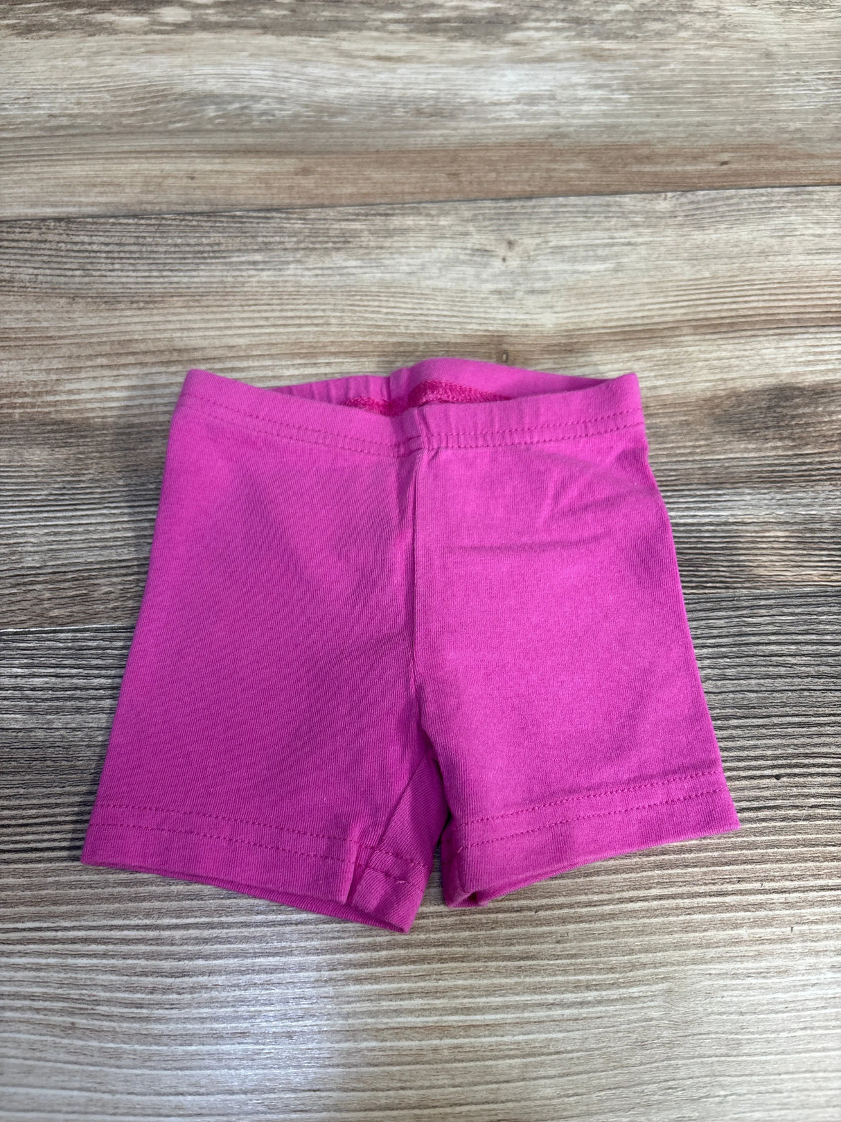 Top-down view of pink baby bike shorts (3 months) showcasing elastic waistband, double-stitched hems, and center seam on wood surface