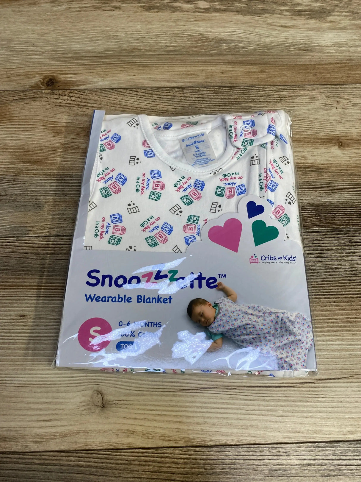 Front view of packaged Cribs for Kids Snoozzzette wearable blanket, size 0–6 months, white with multicolored block print on wood background