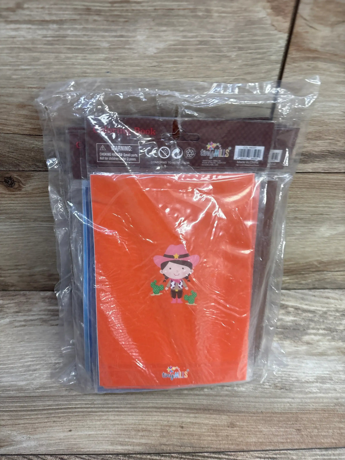 Front view of Tiny Mills Western coloring books and crayons 10 pack showing orange cowgirl cover sealed in plastic on wood background