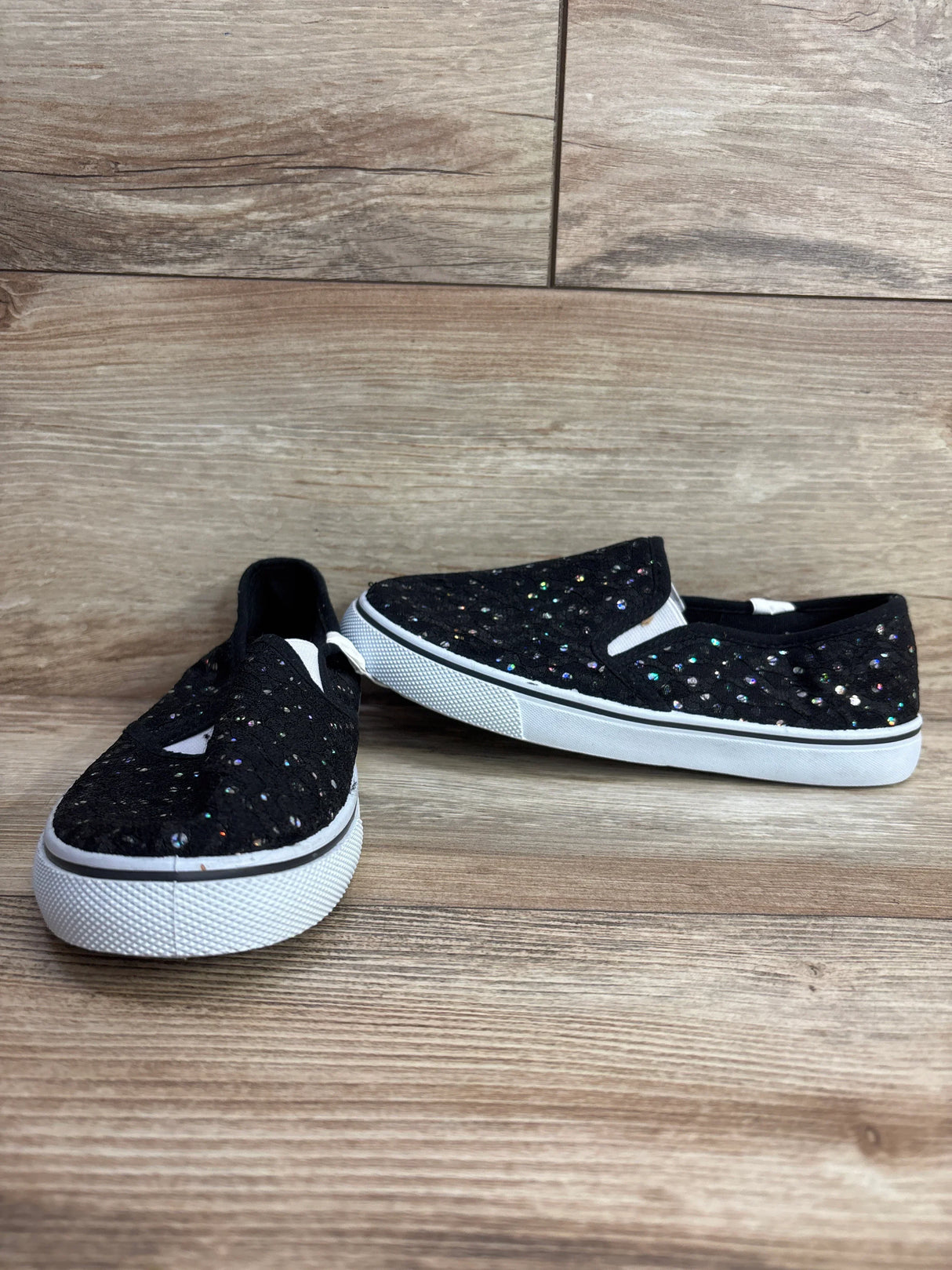 Pair of black kids slip-on shoes with multicolor sparkly sequins and white rubber soles on wooden floor