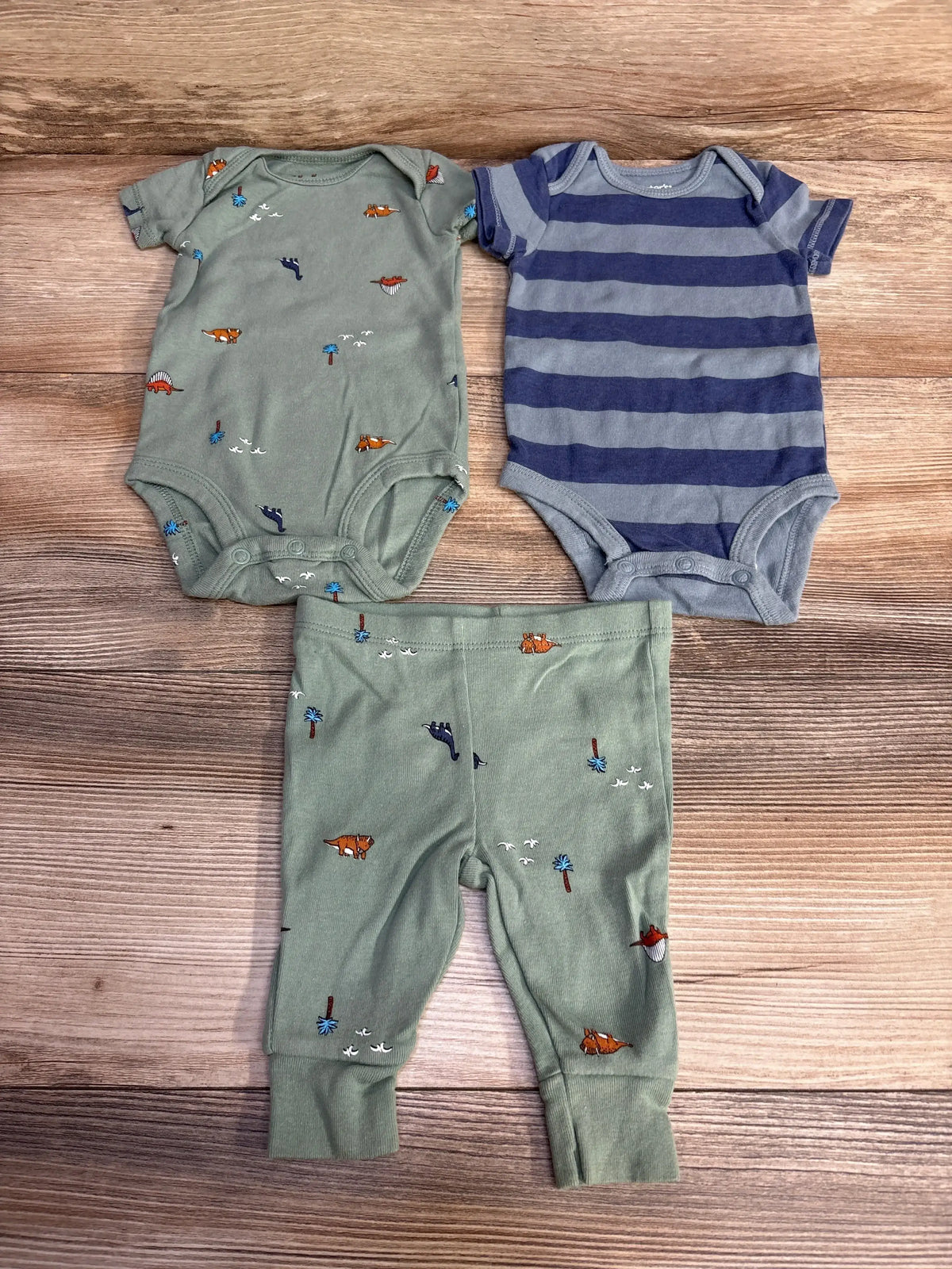 Flat lay of 3-piece baby bodysuit set showing green dinosaur print bodysuit, blue gray striped bodysuit and matching green pants on wood background