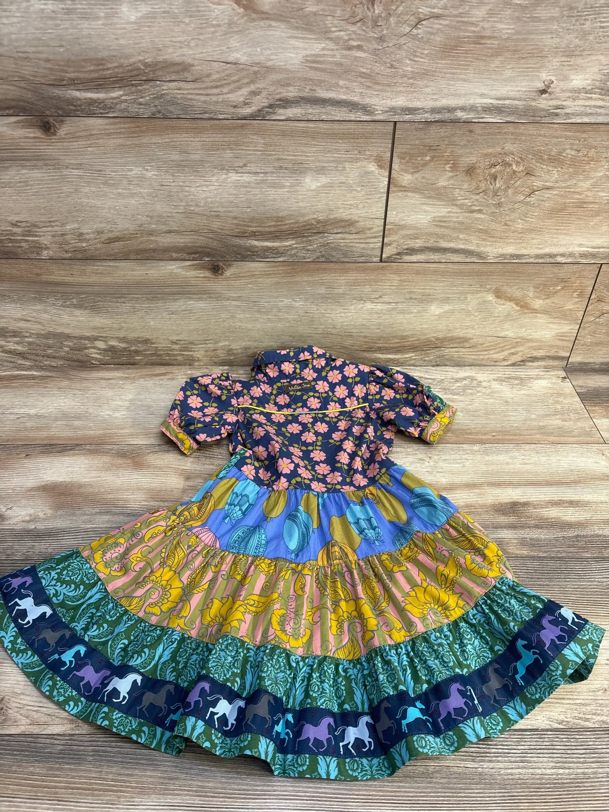 Flat lay of Matilda Jane 2T dress showing navy floral bodice, multicolor tiered skirt with horse-print trim on wood floor background