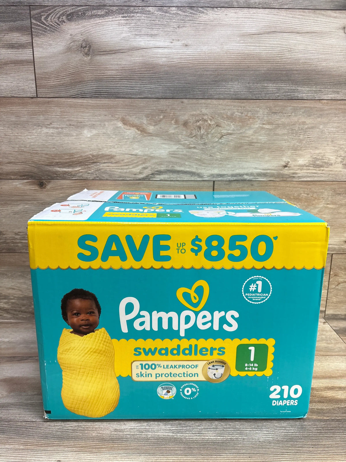 Front view of Pampers Swaddlers diapers size 1 box showcasing 210 diapers with a baby in a yellow swaddle on a wood background