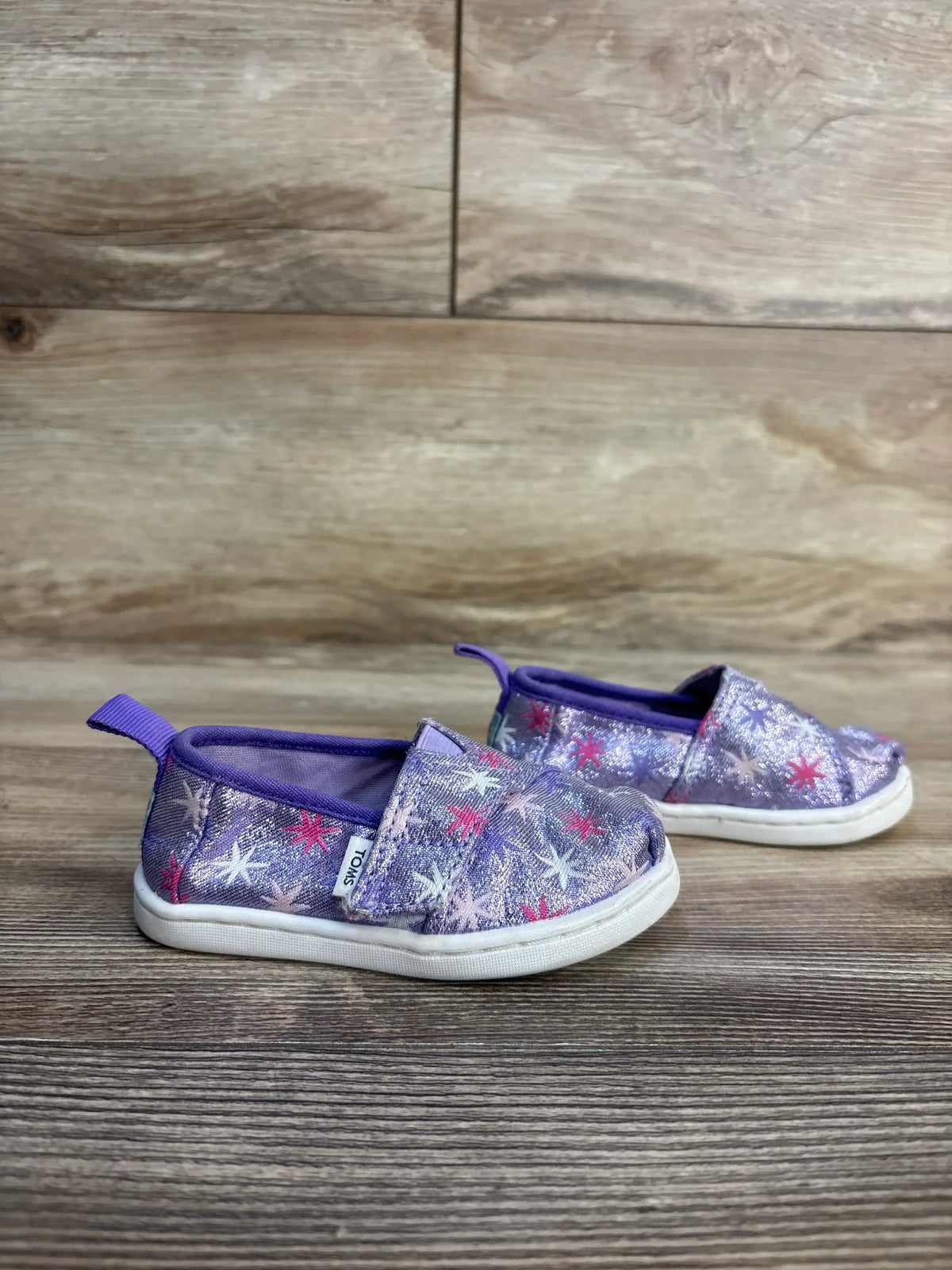 Side view of Toms Tiny Alpargata purple glitter slip-on toddler shoes (size 5C) showcasing star print and white rubber soles on wood floor