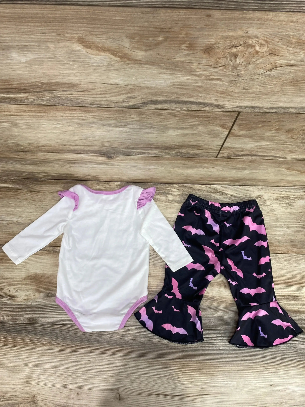 Flat lay of 2-piece baby set: white long sleeve bodysuit with pink trim and black ruffle pants with pink bat print on wood