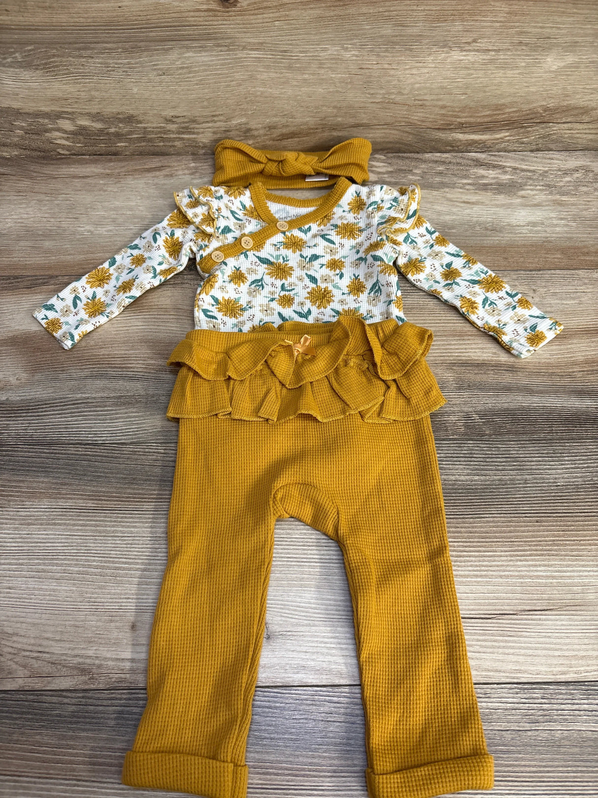 Flat lay of 3pc waffle bodysuit and pants set, mustard floral ruffle top, waffle-knit pants, and matching bow headband, wood floor