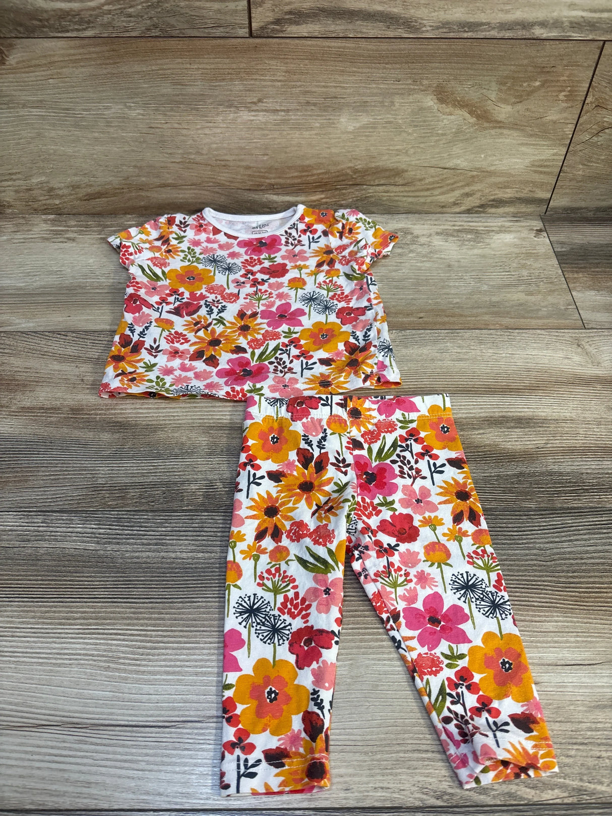 Top view of Fred & Flo newborn 2-piece floral pajama set showcasing short-sleeve crewneck top and matching leggings with pink, orange, and yellow flowers on a wood floor