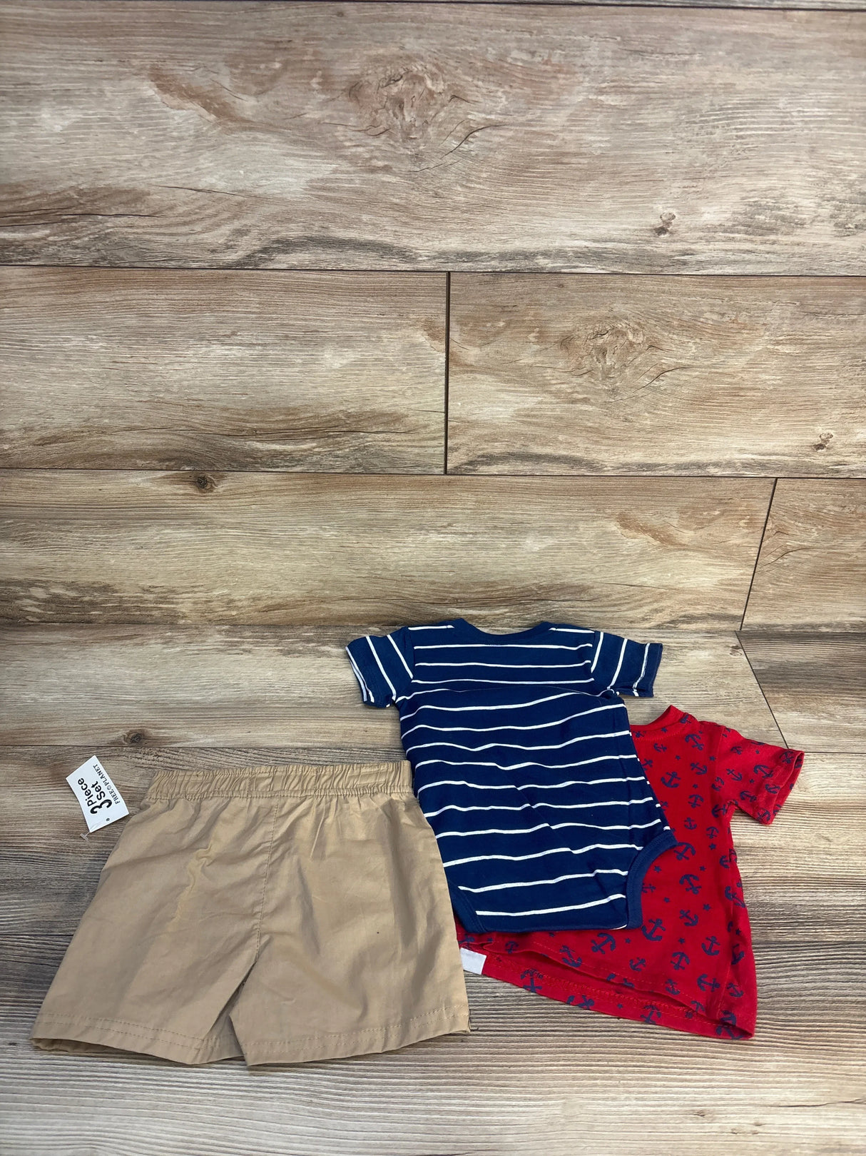Toddler boy's summer outfit set with tan shorts, blue striped bodysuit, and red anchor print bodysuit on wood floor