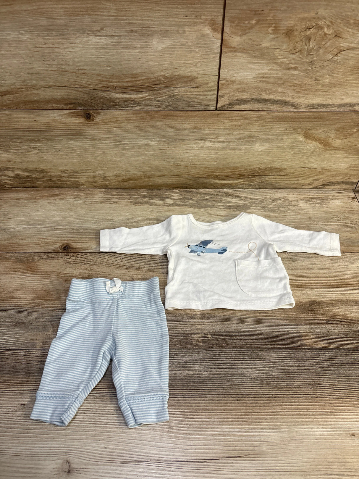 Top-down view of newborn 2-piece set: white long-sleeve shirt with blue airplane and pocket, blue-striped pants on wood
