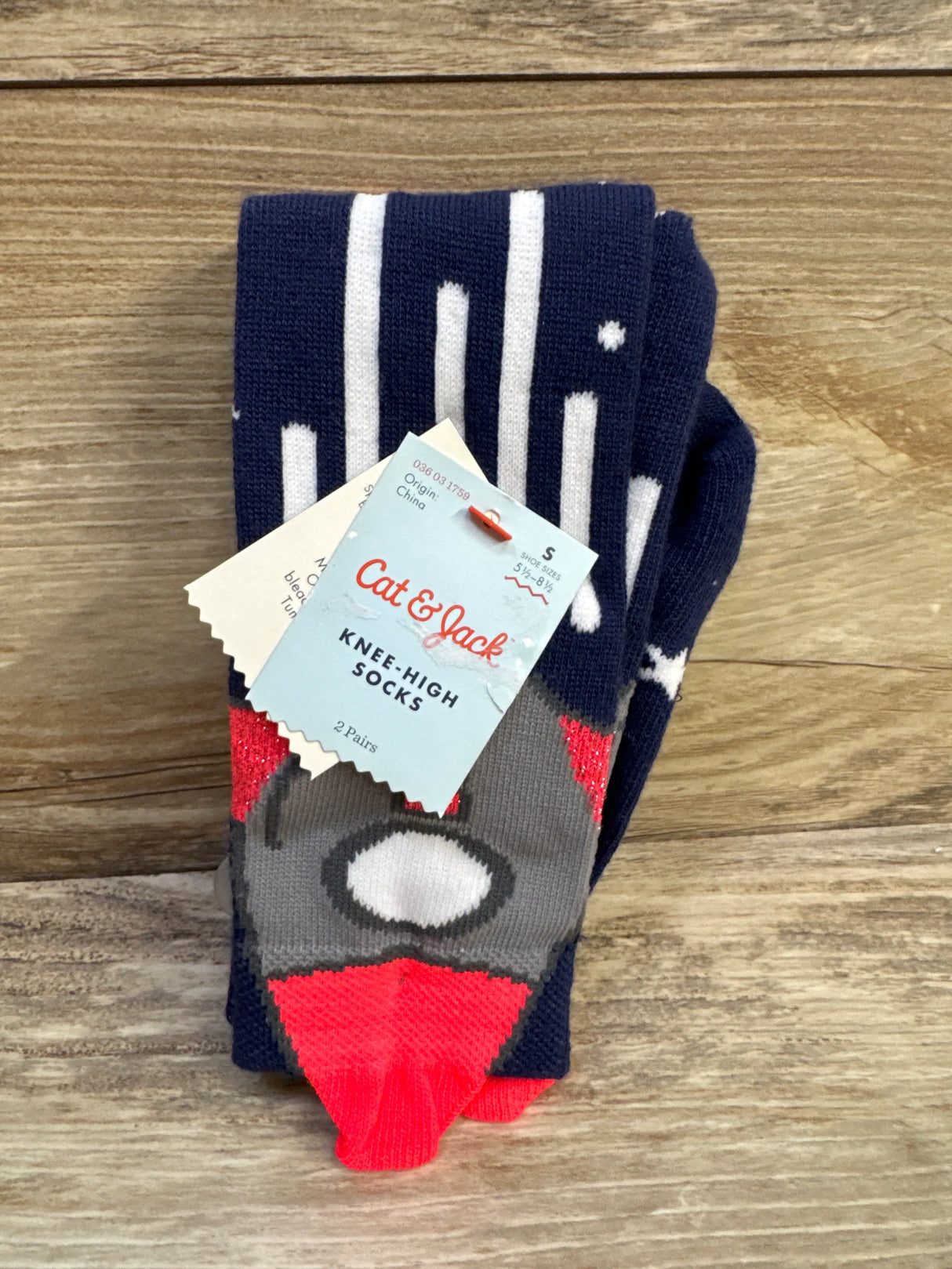 Top view of Cat and Jack knee-high socks showing navy and white stripes, gray rocket face and bright red toes, two-pack with retail tag on wood background