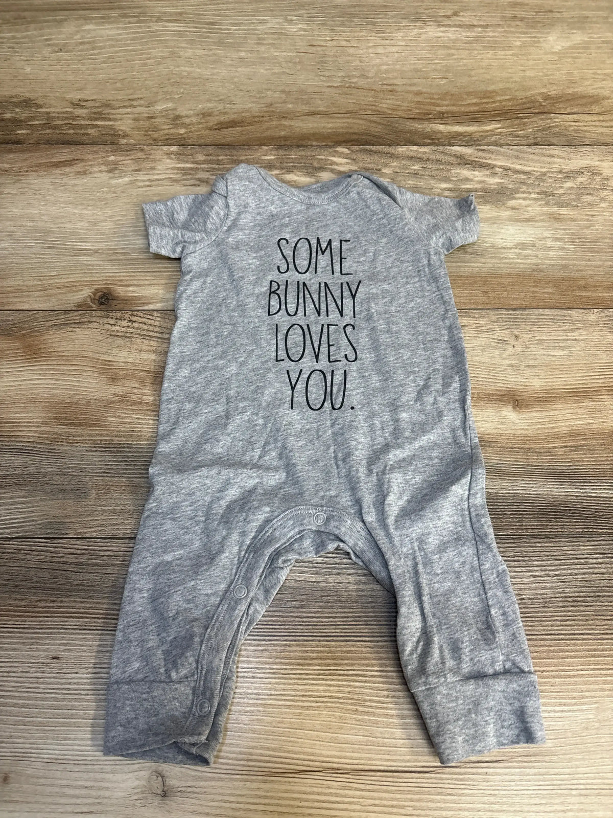 Gray baby onesie with 'Some Bunny Loves You' text, laid on wood floor, ideal for Easter or spring baby outfits.