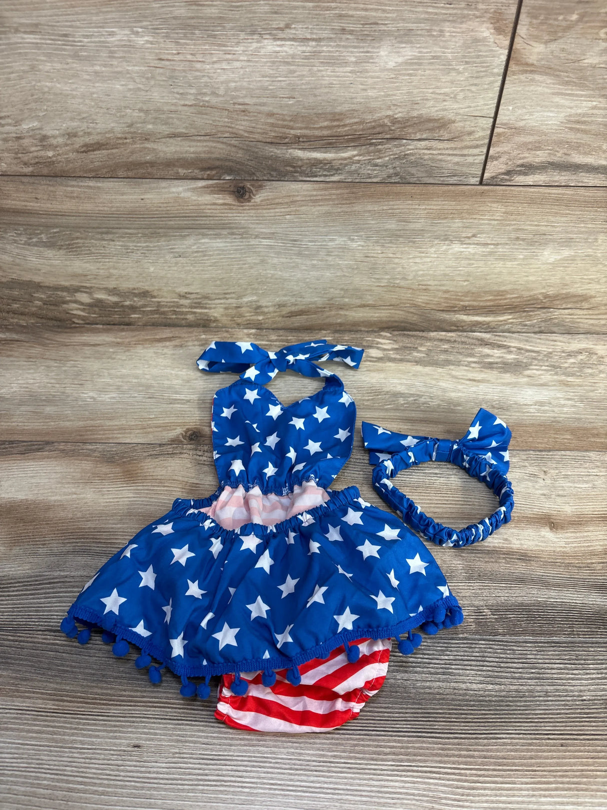 Top down view of halter bubble romper and headband showing blue stars, striped bloomers, and pom pom trim on wood background