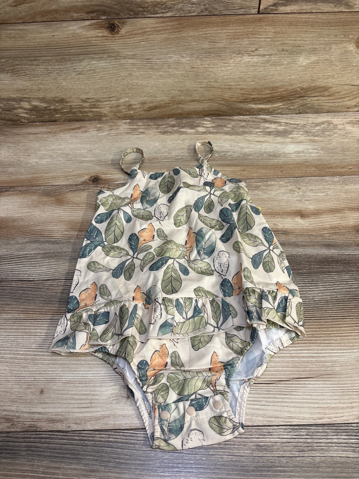 Top-down view of beige leaf-print skirted bubble romper (size 18–24m) showing ruffled skirt overlay and snap crotch on wood background