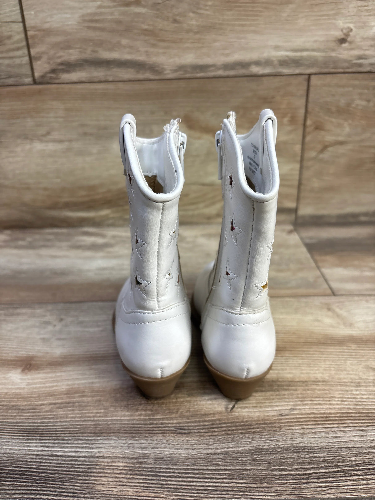 Back view of white toddler cowgirl boots showing star cutouts on shaft, inner zippers, and tan stacked heels on wood floor