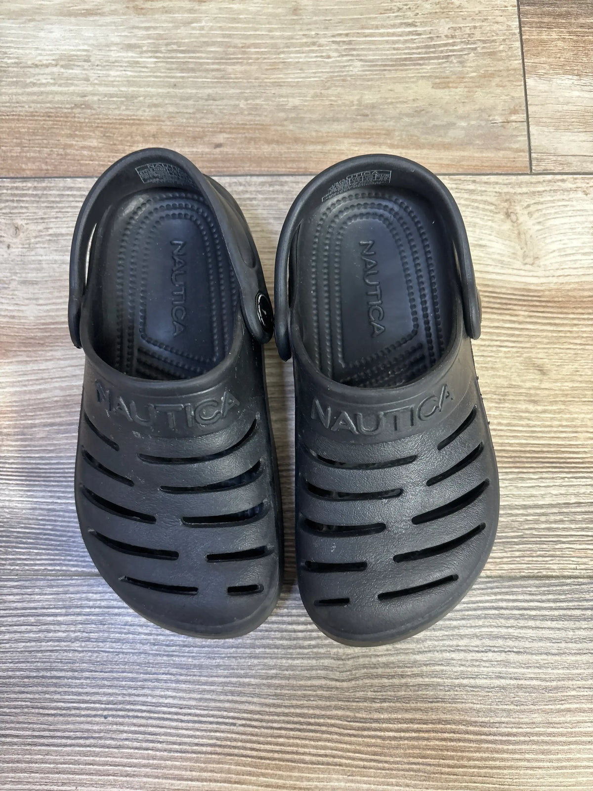 Top-down view of black Nautica River Edge toddler clogs showing ventilation slits, heel straps, and embossed Nautica logo on wood floor