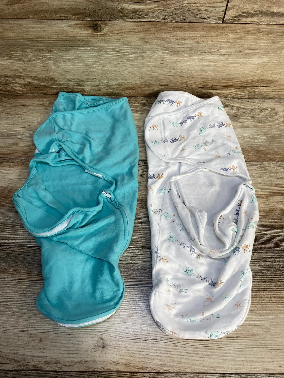 Top-down view of Aden and Anais 2-pack newborn swaddle wraps, solid teal and white animal-print, on wood floor