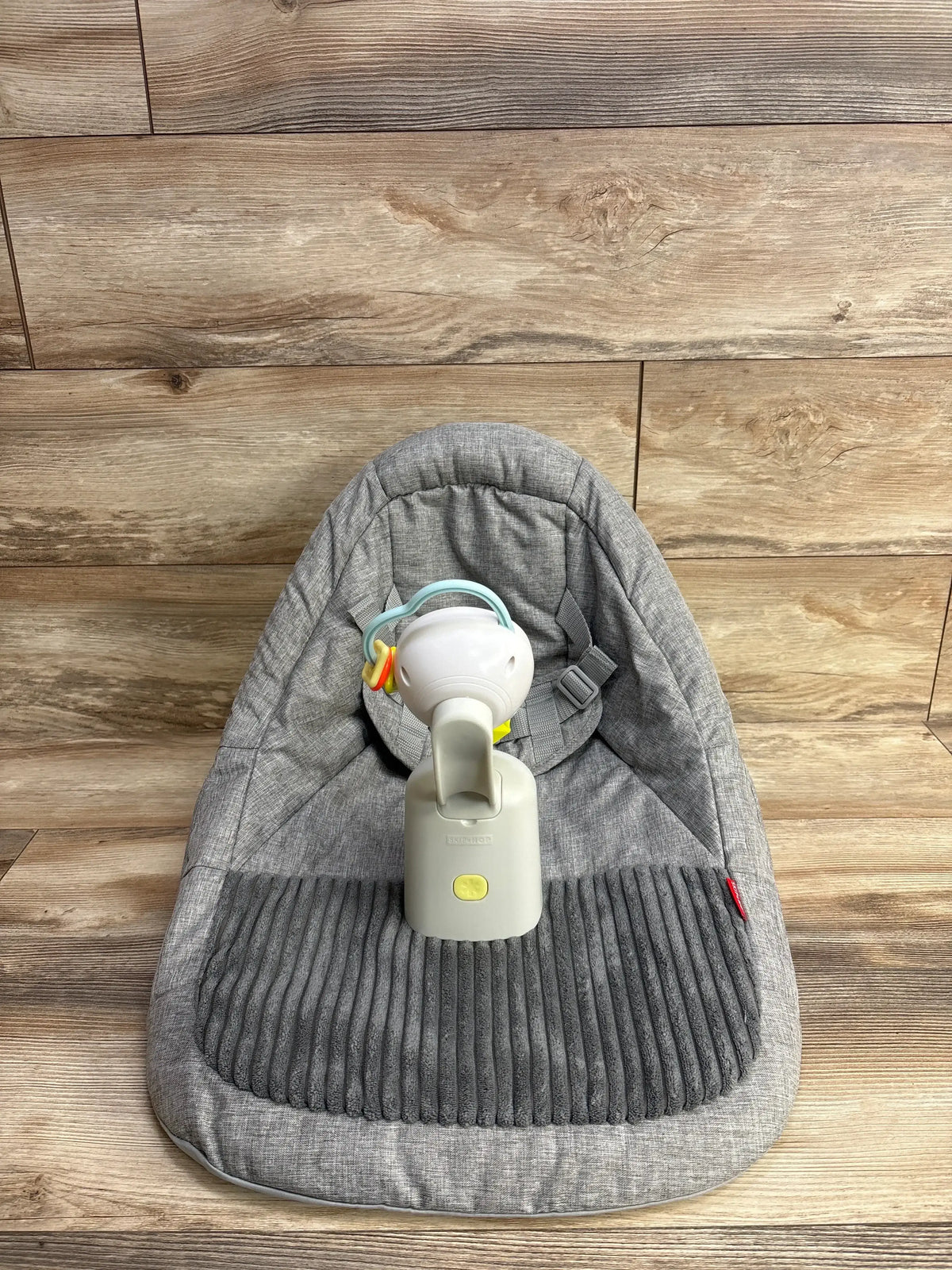 Front view of Skip Hop Silver Lining Cloud upright floor seat showing gray padded ribbed cushion, safety harness, and attached white toy on wooden floor