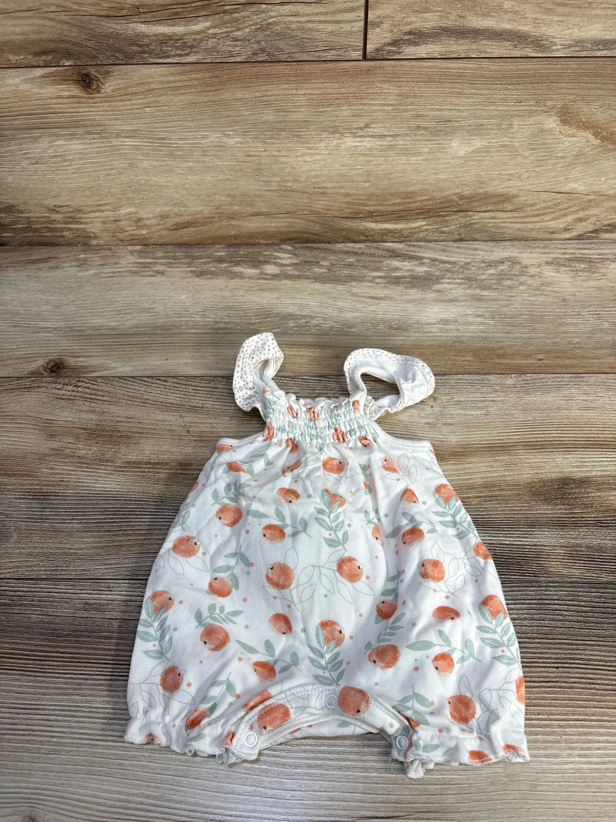 Front view of white Rabbit + Bear shortie romper with orange fruit and green leaf print and snap closures on wood background