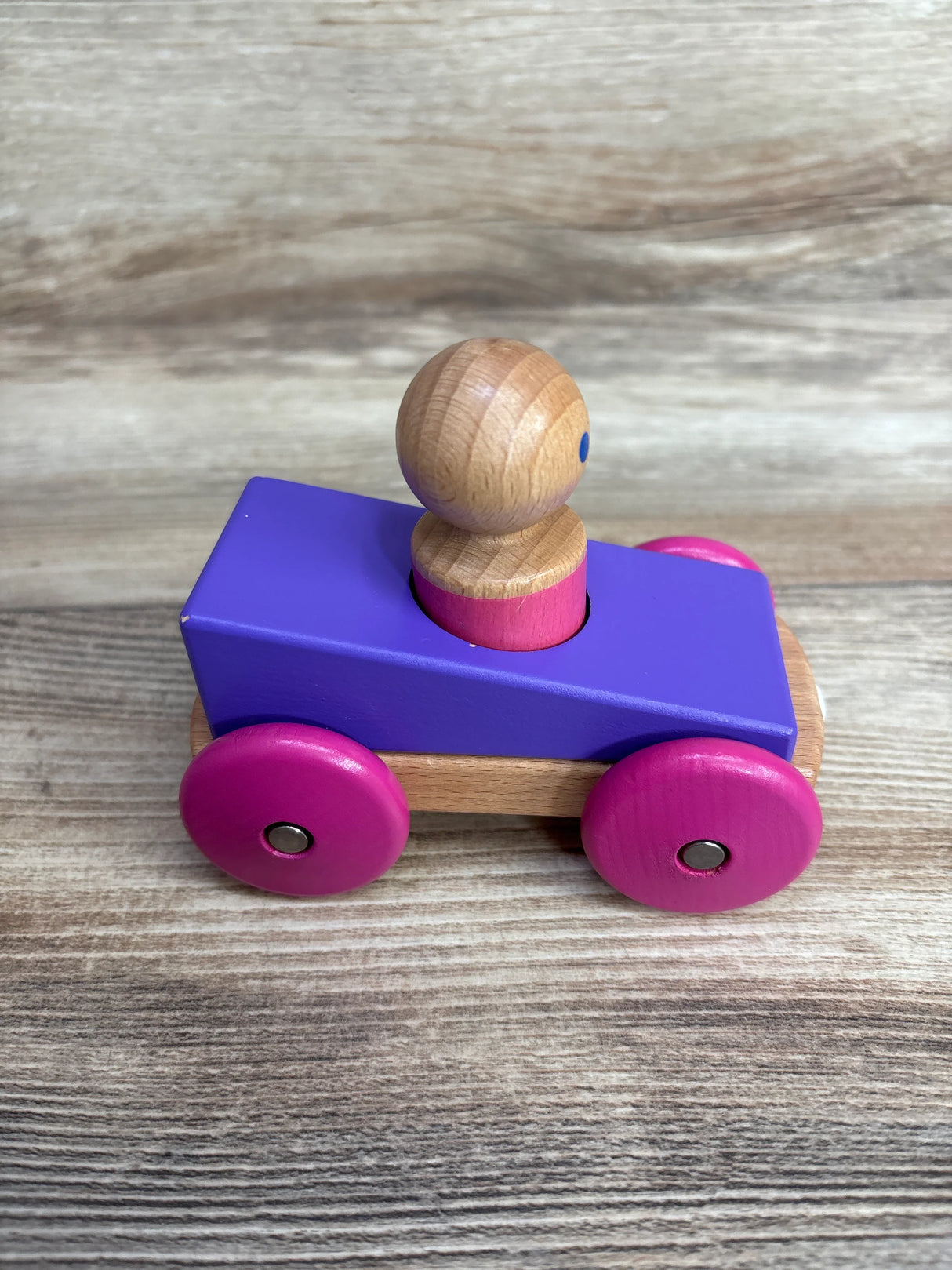 Wooden toy car with purple body, pink wheels, and natural wood peg figure on a wooden surface