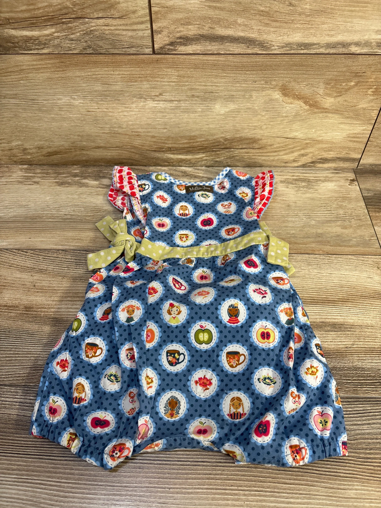 Front flat lay of Matilda Jane Teachers Pet romper, blue polka-dot with round character medallions, green polka-dot waist tie and ruffle sleeves on wood background