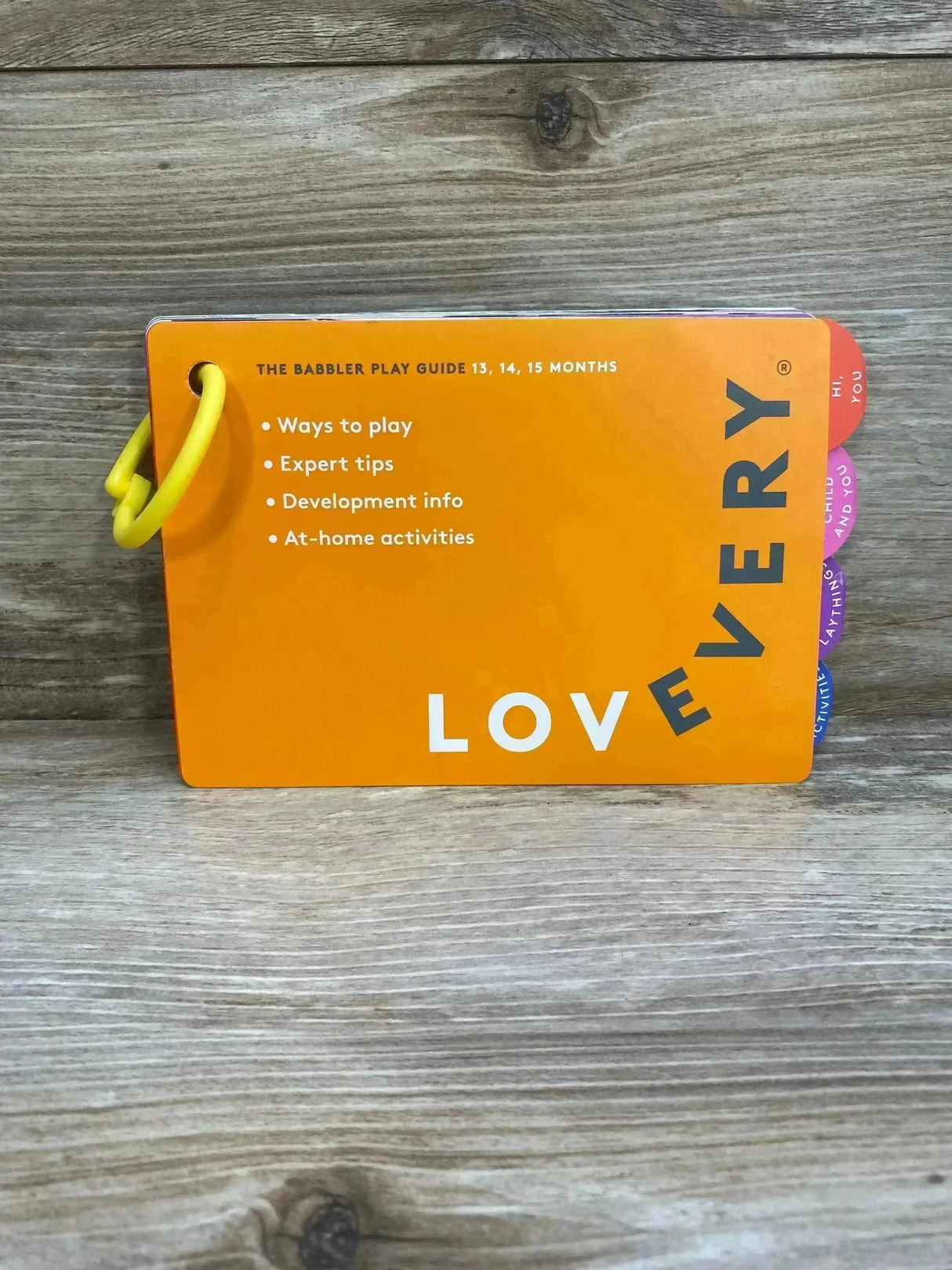 Front view of Lovevery The Babbler play guide 13-15 months showcasing orange cover, yellow plastic ring, and colored tabs on wood background