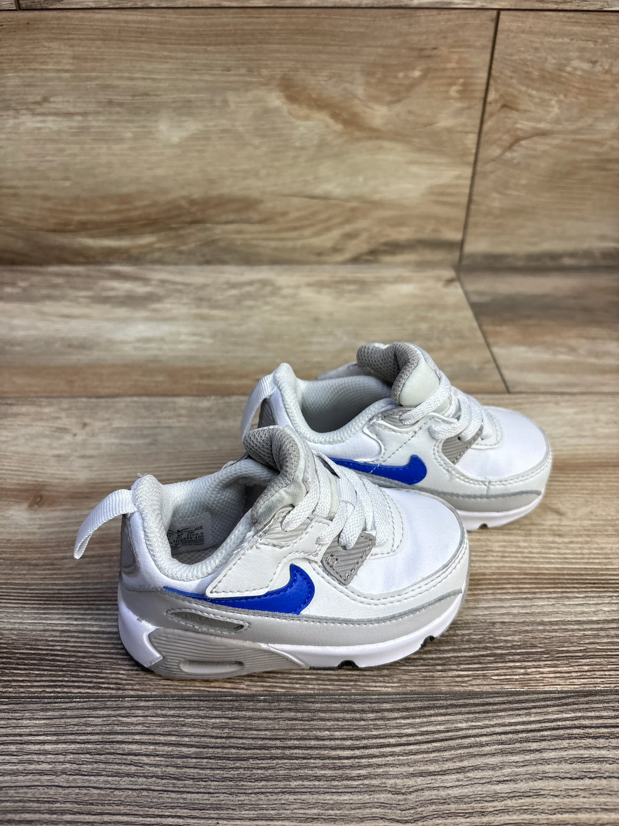 Angled view of toddler Nike Air Max 90 EasyOn sneakers with blue Swoosh, gray trim and pull tab on wood background