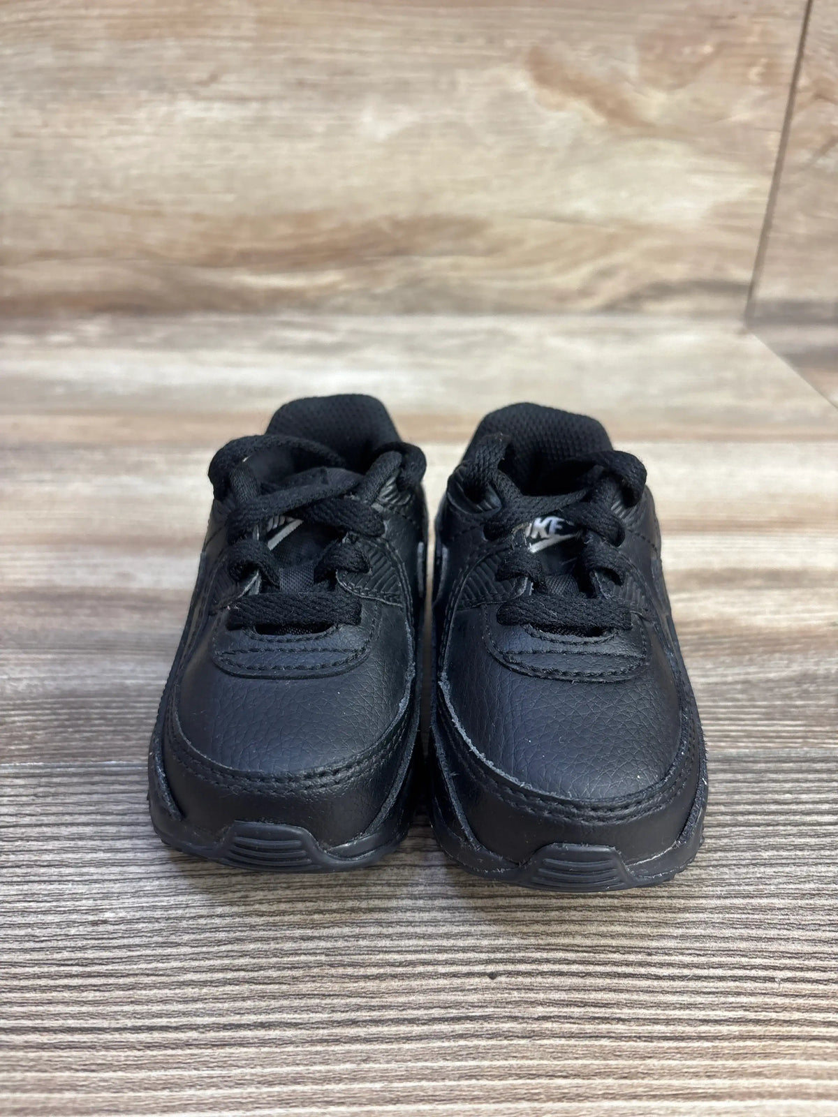 Front view of Nike Air Max 90 TD Triple Black sneakers showcasing black textured uppers and laces on a wood floor