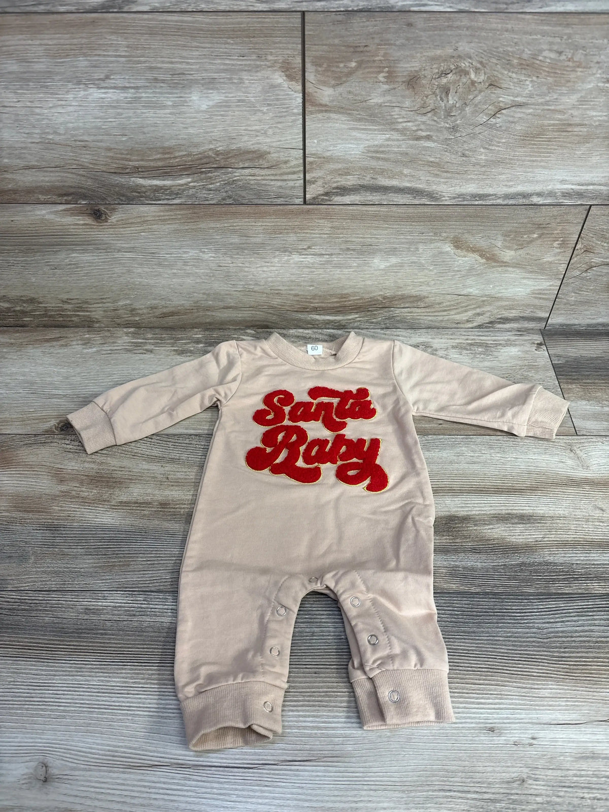 Beige baby onesie with red 'Santa Baby' embroidery, long sleeves, snap closures, laid on wood floor