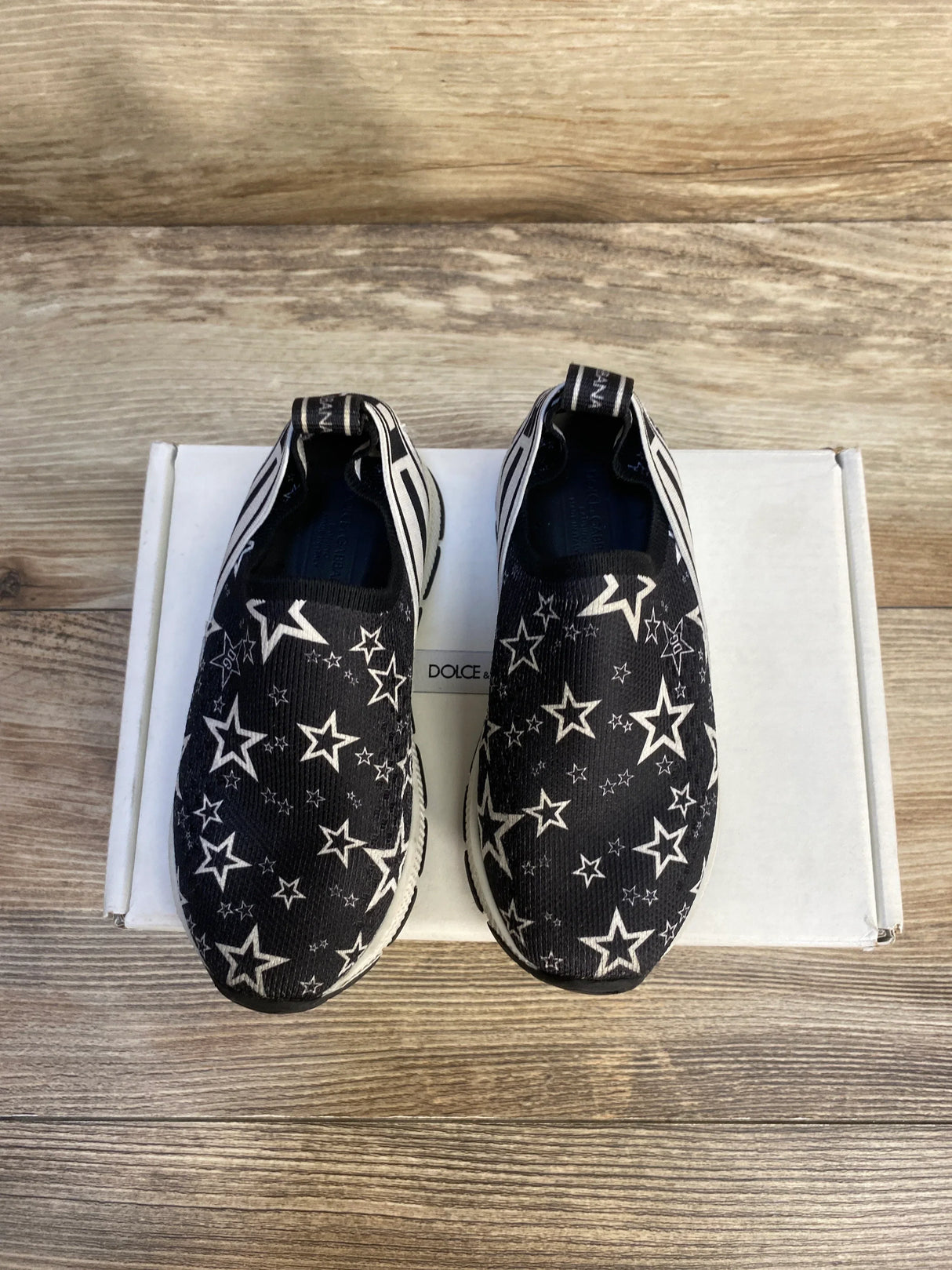 Black slip-on kids shoes with white star pattern on box, wooden background