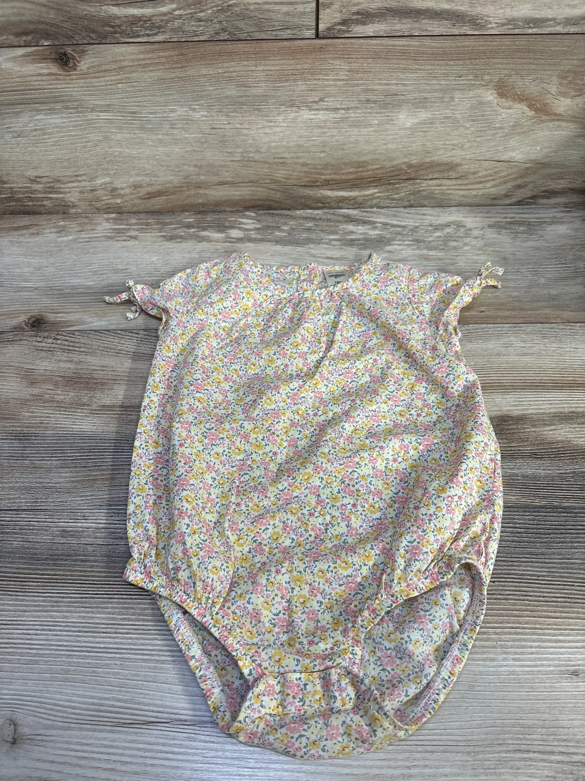 Flat lay of OshKosh yellow floral bubble romper, size 18m, with cap sleeves, ties, and elastic leg openings on wood