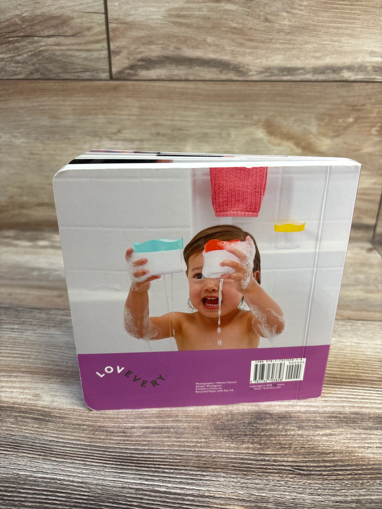 Back of Lovevery Bedtime for Zoe board book showing toddler in bubbly bath pouring cups, purple strip on wood surface
