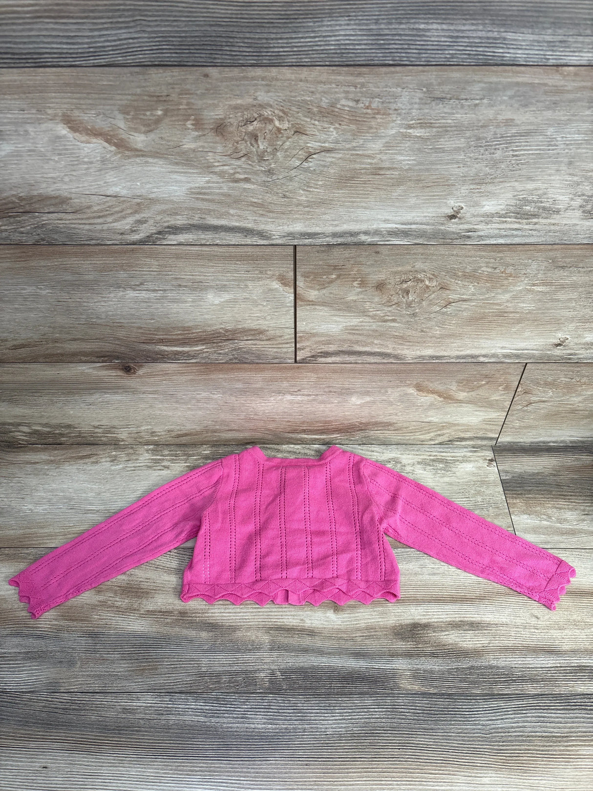 Pink knitted long sleeve baby cardigan with scalloped edges on rustic wooden floor