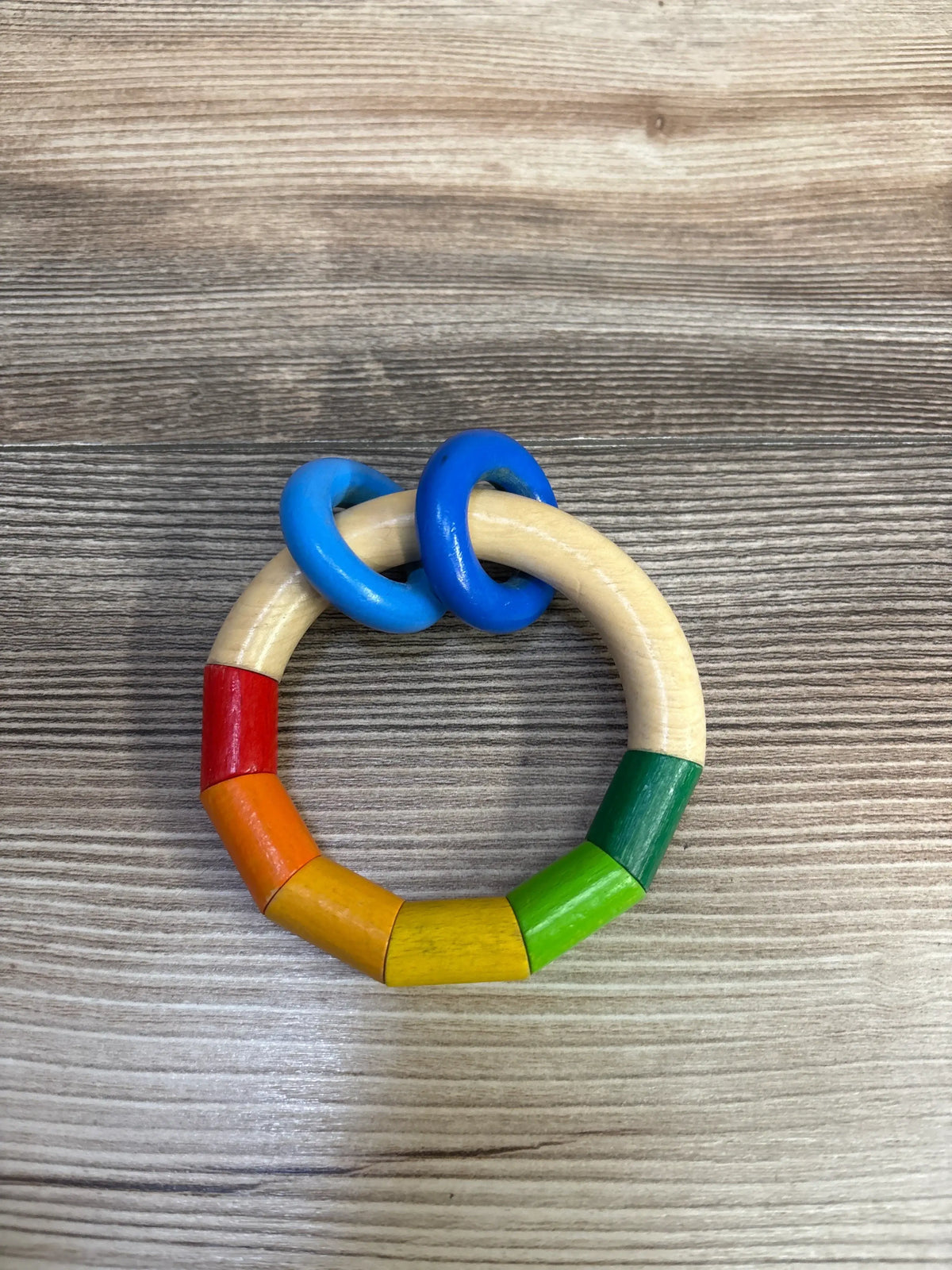 Top view of HABA Kringelring Wooden Clutching Toy showcasing rainbow-colored beechwood beads and two blue plastic rings on a wood-grain background