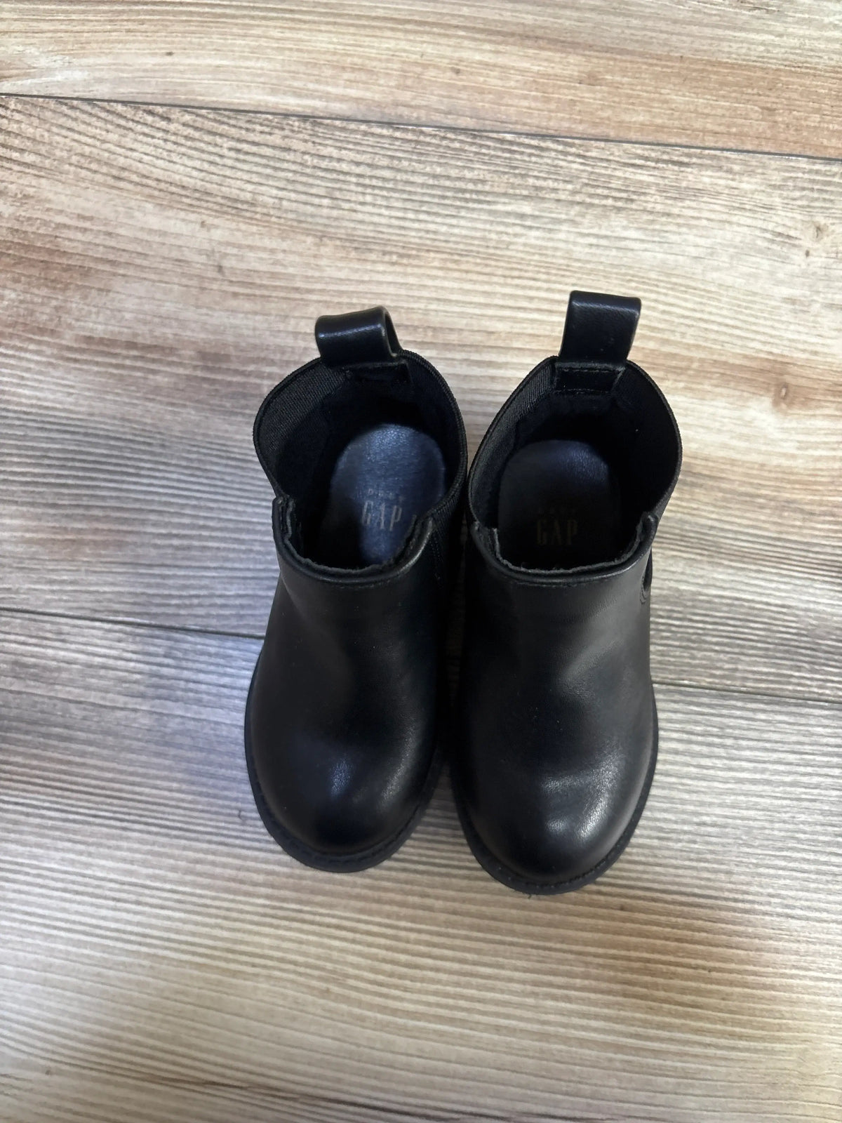 Top-down view of Baby Gap black leather ankle boots showing rounded toes, pull tabs, elastic side panels on light wood floor