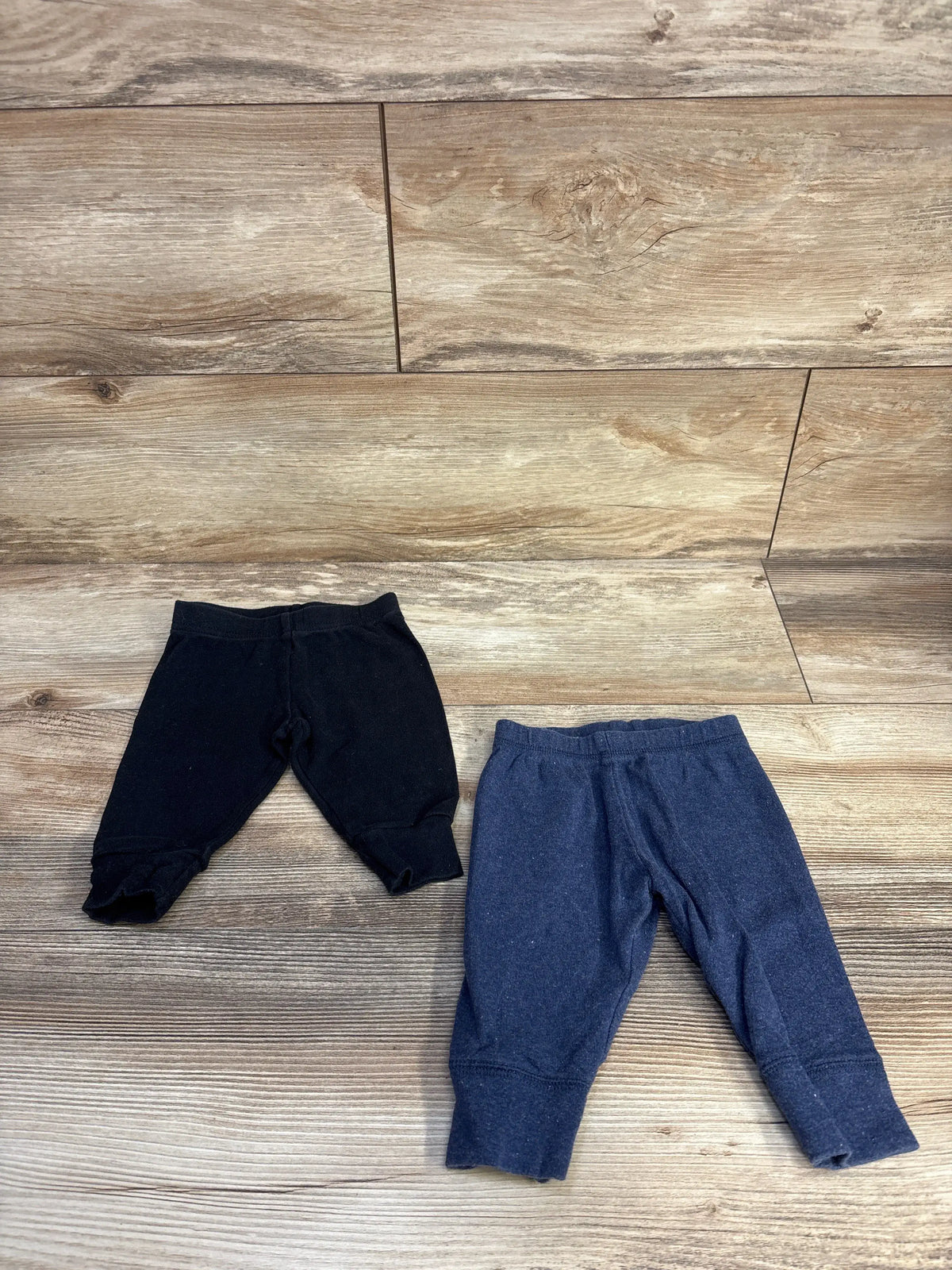 Two pairs of Simple Joys baby pants in black and navy blue on a wood floor.
