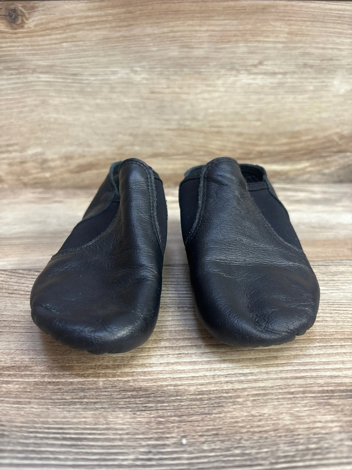 Front view of Capezio girls’ black leather jazz slip-on shoes showing elastic sides and light scuffing on wooden floor